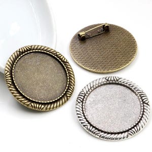 Puede incluir: Tres blancos redondos de metal para la elaboración de joyas. Dos son de bronce con un borde de cuerda y uno es de plata con un borde de cuerda. Los blancos de bronce tienen un broche de seguridad.