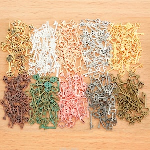 May include: Assortment of antique-style key charms in various colors, including gold, silver, bronze, green, and pink. The keys are arranged in rows on a wooden surface.