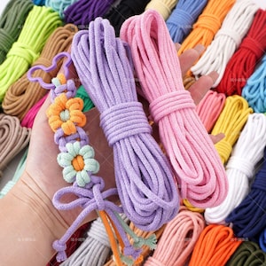 May include: A hand holds bundles of colorful cotton rope in shades of purple, pink, and other colors. A macrame bracelet with orange and green flower details is also visible. The background features more bundles of rope in various colors.