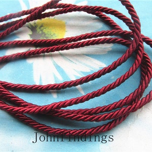 May include: Burgundy red twisted cord, a type of decorative trim, is shown in a close-up image. The cord is made of a soft, silky material and has a subtle sheen.