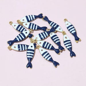 May include: A selection of fish-shaped charms with a nautical theme. Each charm features a white body with navy blue stripes and a navy blue tail. The charms have a gold-coloured loop at the top for attaching to jewellery or other crafts.