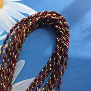 May include: A close-up of a red and gold braided cord. The cord is made of multiple strands of yarn twisted together.