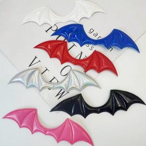 May include: Six bat wing-shaped patches in various colors including white, blue, red, iridescent silver, black, and pink. Each patch features a distinct pointed wing design with raised structural lines, arranged in a scattered pattern on a white surface.