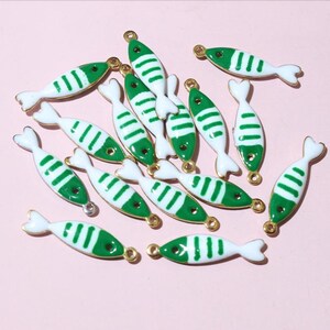 May include: A selection of fish-shaped charms. Each charm is white with green stripes and a gold-coloured border. The charms have a small loop at the top for attaching to jewellery or other crafts. The background is a solid, light pink colour.