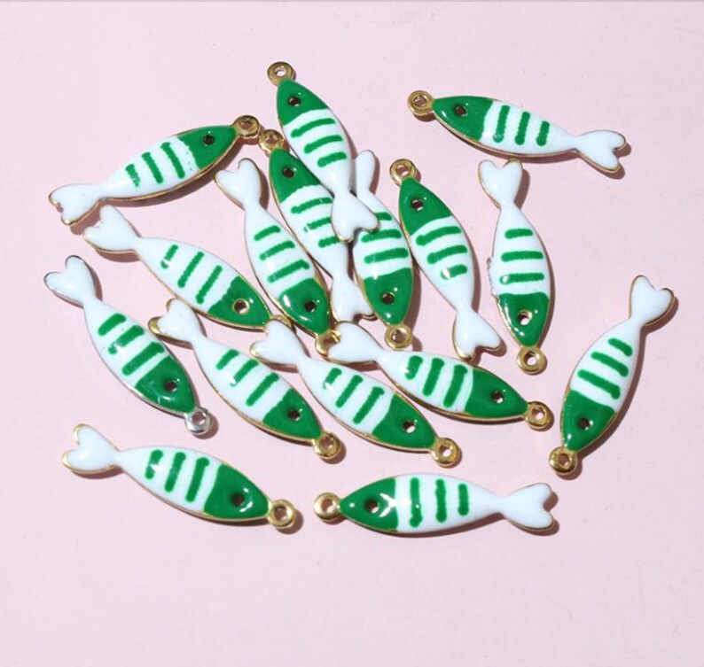 May include: A selection of fish-shaped charms. Each charm is white with green stripes and a gold-coloured border. The charms have a small loop at the top for attaching to jewellery or other crafts. The background is a solid, light pink colour.