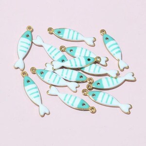 May include: A selection of fish-shaped charms. Each charm is white with turquoise stripes and a turquoise head, edged in gold. The charms have a small loop at the top for attaching to jewellery or other crafts. The background is a pale pink.
