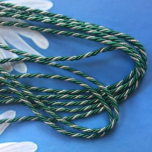 May include: Close-up of green and silver twisted cord. The cord is a vibrant green with a metallic silver thread twisted throughout. The cord is arranged in loose loops against a blue background.