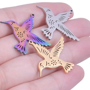 May include: Three hummingbird pendants in different metallic finishes. One is iridescent with rainbow hues, another is silver, and the third is gold. Each pendant features a cutout design of a hummingbird in flight, with intricate wing and tail details.
