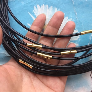 Puede incluir: Un conjunto de cordones de cuero negro con cierres de metal dorado. Los cordones están apilados uno encima del otro y se sostienen en una mano.