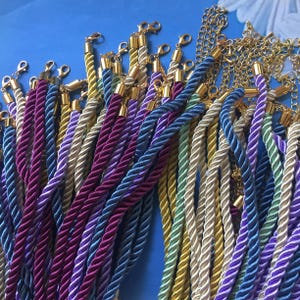 Puede incluir: Una colección de coloridos cordones de cuerda trenzada con cierres y detalles de cadena de metal dorado. Los cordones son en tonos de morado, azul, verde y dorado, creando una exhibición vibrante. Los cordones son probablemente para joyería o proyectos de artesanía.