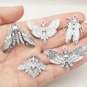 Puede incluir: Cinco dijes plateados con forma de polilla con detalles intrincados. Cada dije presenta un diseño de polilla con un motivo de calavera en la cabeza. Los dijes varían en forma y diseño de alas, ideales para manualidades.