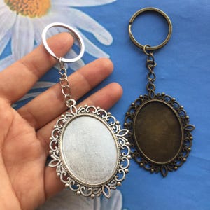 Puede incluir: Dos llaveros ovalados ornamentados. Uno es plateado, el otro es bronce. Ambos tienen un borde de filigrana decorativo y un espacio ovalado en blanco para una foto o diseño. Cada llavero tiene un anillo y una cadena.