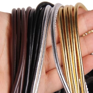 May include: A close-up of a selection of cords in various colours and textures, including brown, black, silver, and gold. The cords are arranged in a fan shape and are likely used for crafting or jewellery making.