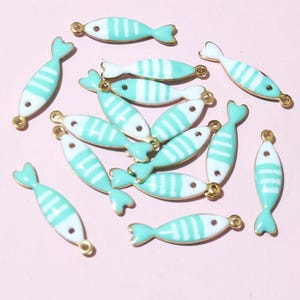 May include: A selection of small, fish-shaped charms. Each charm is white with turquoise stripes and a turquoise tail fin. The charms have gold-coloured loops for attaching to jewellery or other crafts. The background is a light pink colour.