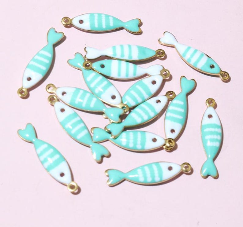 May include: A selection of small, fish-shaped charms. Each charm is white with turquoise stripes and a turquoise tail fin. The charms have gold-coloured loops for attaching to jewellery or other crafts. The background is a light pink colour.