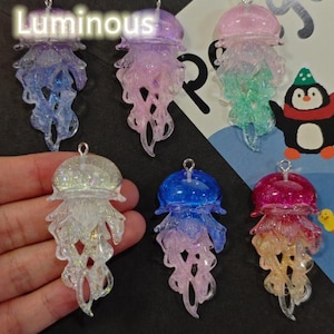 Peut inclure: Six pendentifs méduse translucides avec des détails scintillants et une finition lumineuse. La collection comprend des modèles bleus, violets, roses, transparents et multicolores, chacun avec une petite boucle métallique pour les suspendre.