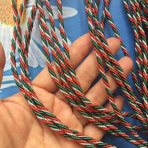 May include: Close-up of several strands of twisted cord in red, green, and silver. The cords are tightly wound together, creating a textured appearance. The cords are likely used for crafting or decorative purposes.