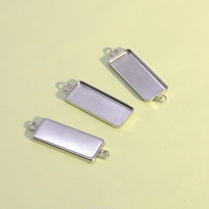 Puede incluir: Tres bases rectangulares para joyería en metal plateado con un centro liso y hundido, que presentan un pequeño aro circular en cada uno de los dos extremos cortos, diseñadas para sujetar inserciones o elementos decorativos.