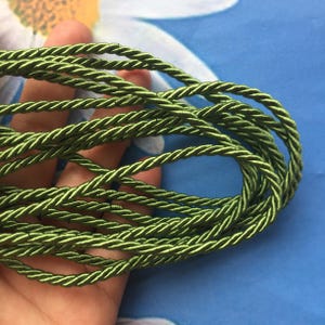 May include: Close-up of a coil of green twisted rope. The rope is a vibrant shade of green, with a slight sheen. The rope is tightly wound, creating a textured surface. The background is a blurred blue and white floral pattern.