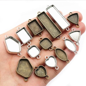 May include: A collection of antique bronze and silver-toned bezel trays in various shapes, including rectangular, square, and heart. Each bezel has a loop for attaching to jewelry. The trays are empty, ready for resin or other embellishments.