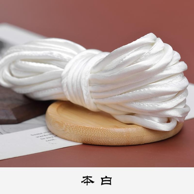 May include: A close-up of a white satin cord, coiled and displayed on a wooden coaster. The text "本白" is visible below the cord.