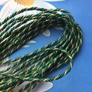 May include: A close-up of a spool of green and gold twisted cord. The cord is made of a soft, silky material and has a subtle sheen.