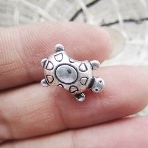 May include: A silver charm in the shape of a turtle. The turtle has a detailed shell with a pattern of circles and a small head with a mouth.