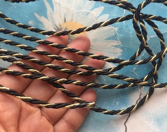 May include: Black and gold twisted cord trim. The cord is made of a black fabric with a gold thread running through it. The cord is shown in a hand.