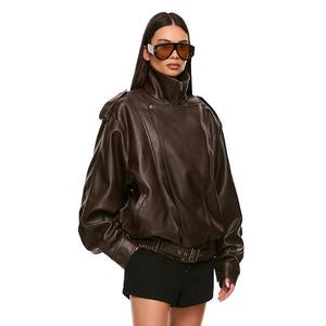 Könnte beinhalten: Braune Lederjacke mit hohem Kragen und Gürtel, kombiniert mit schwarzen Shorts und kniehohen braunen Stiefeln. Die Stiefel haben einen kleinen Absatz und eine dekorative Schnalle. Die Person trägt eine Sonnenbrille.