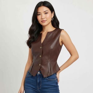 Puede incluir: Un chaleco de cuero marrón con cuello en V y tres botones dorados. El chaleco tiene un corte entallado con pliegues en la cintura. La modelo lleva vaqueros azules.