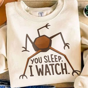 May include: Cream-colored sweatshirt with a brown insect-like graphic and the text "YOU SLEEP. I WATCH." The graphic features a central brown shape with long, thin legs and arms. The sweatshirt is folded, with a glimpse of denim jeans visible.