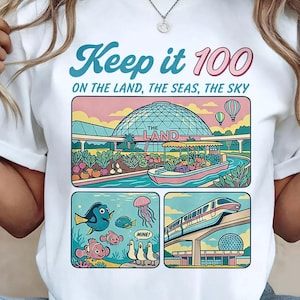 May include: White t-shirt with a colorful graphic featuring the text "Keep it 100" and illustrations of a theme park. The design includes images of a boat ride, fish, and a monorail.