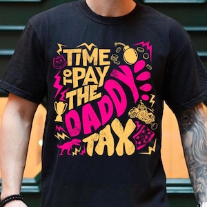 May include: A black short-sleeved t-shirt featuring a graphic print in yellow and hot pink. The text reads "TIME TO PAY THE DADDY TAX" surrounded by illustrations of a bomb, a trophy, a motorcycle, a dinosaur, and various geometric shapes.