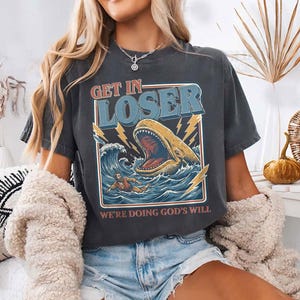 Puede incluir: Camiseta gris oscuro con un dise&ntilde;o gr&aacute;fico que presenta una ballena con la boca abierta en el oc&eacute;ano, rayos y el texto "GET IN LOSER" y "WE'RE DOING GOD'S WILL". La camiseta tiene un aspecto vintage.