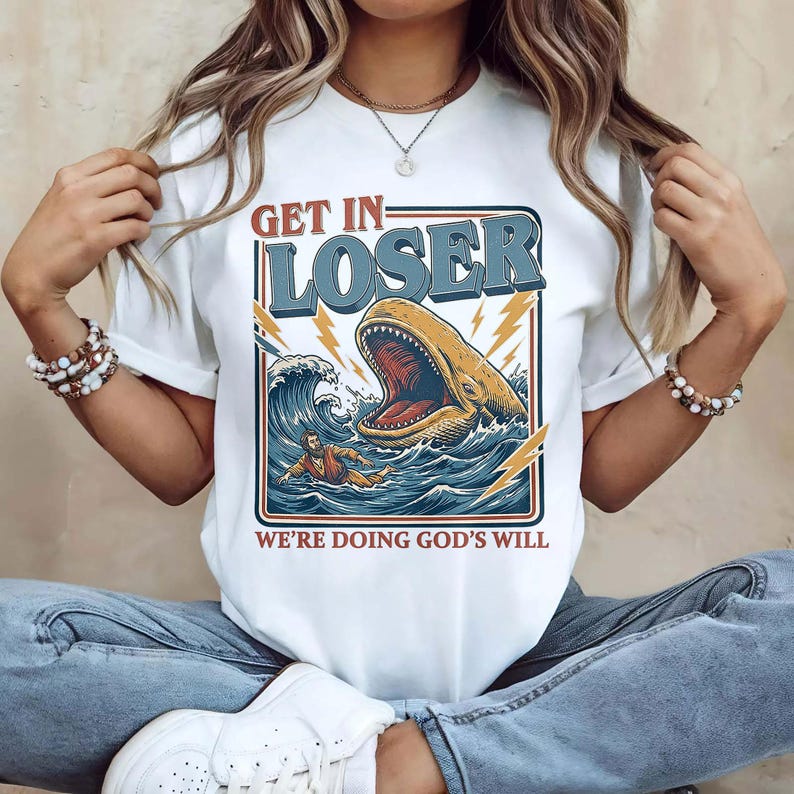 Puede incluir: Camiseta blanca con un dise&ntilde;o gr&aacute;fico que presenta una ballena con la boca abierta en el oc&eacute;ano, una persona en el agua y el texto "GET IN - LOSER WE'RE DOING GOD'S WILL" en rojo y azul.