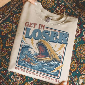 Puede incluir: Sudadera beige con un dise&ntilde;o gr&aacute;fico que presenta una ballena con la boca abierta, olas y una persona en el agua. El texto dice "GET IN LOSER" y "WE'RE DOING GOD'S WILL" en un estilo vintage.