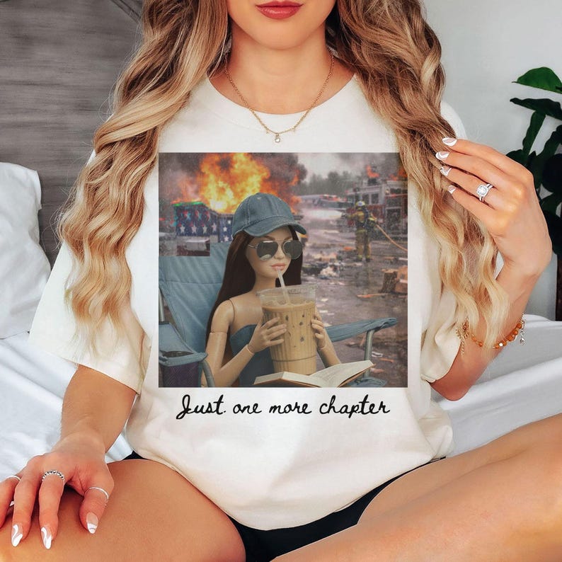 May include: Cream-colored t-shirt featuring a graphic of a doll with sunglasses, a hat, and an iced coffee, sitting in a chair with a book. The background shows a burning building. The text "Just one more chapter" is below the graphic.