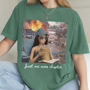 May include: Green t-shirt featuring a graphic of a doll with sunglasses, a drink, and a book, set against a backdrop of a fire. The text "Just one more chapter" is written in cursive. The shirt is a solid green color.