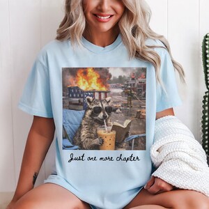 May include: Light blue t-shirt featuring a raccoon wearing sunglasses, drinking a beverage, and reading a book while a fire rages in the background. The text "Just one more chapter" is printed below the image.