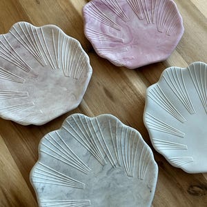 Könnte beinhalten: Vier muschelförmige Dekoschalen in Rosa-, Grau- und Weißtönen. Jede Schale hat ein strukturiertes, fächerartiges Design, das von der Mitte ausgeht. Die Schalen sind auf einer Holzoberfläche angeordnet und zeigen ihre verschiedenen Farben und Muster.