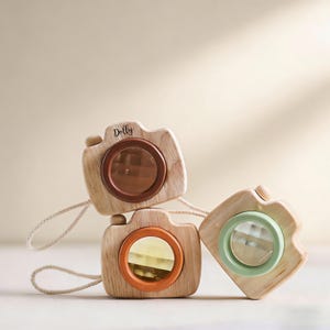 Puede incluir: Tres cámaras de juguete de madera con lentes de diferentes colores: marrón, naranja y verde. Cada cámara tiene un cuerpo de madera clara, una lente y una pequeña correa. La palabra "Dolly" está escrita en una de las cámaras.