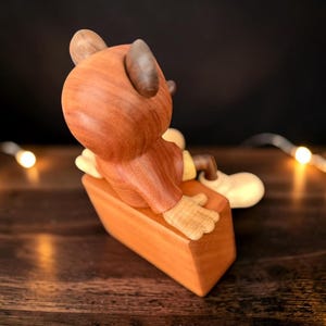 May include: A wooden figurine of a cartoon character, with a round head and large ears, in a seated position. The figure is mainly brown with yellow and cream accents. It is sitting on a rectangular wooden base, with a warm, natural finish.