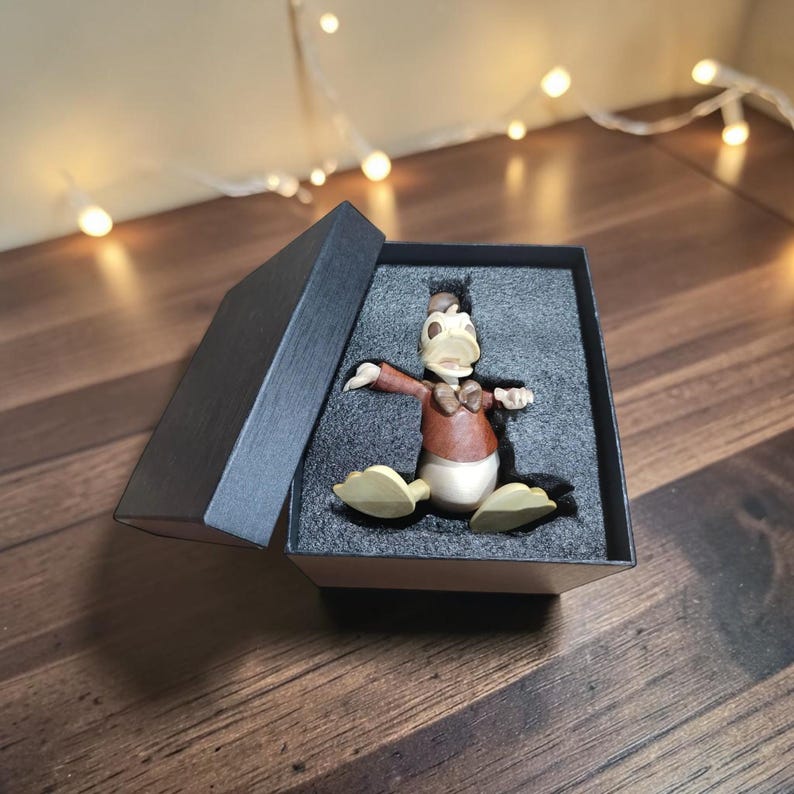 May include: A Donald Duck figurine in a black gift box. The figurine is wearing a brown jacket, bow tie, and hat, with a white belly and yellow feet. The box is open, revealing the figure on a gray surface.