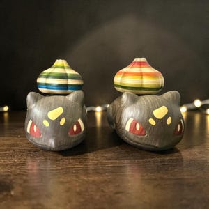 Op de afbeelding: Twee donkergrijze katvormige beeldjes met gele en rode accenten, bekroond met kleurrijke gestreepte houten bolvormen. De beeldjes staan op een houten oppervlak tegen een donkere achtergrond.
