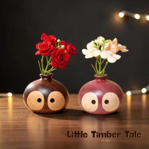 May include: Two wooden vases with an owl-like design, each holding flowers. One vase is dark brown with red roses, the other is purple with white and peach flowers. The text "Little Timber Tale" is at the bottom.