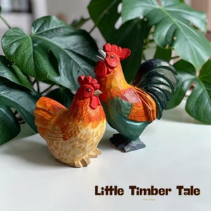 May include: Two hand-carved wooden rooster figurines. One is a hen, the other a rooster, both with red combs and beaks, and orange, yellow, and green plumage. The text "Little Timber Tale" is at the bottom.