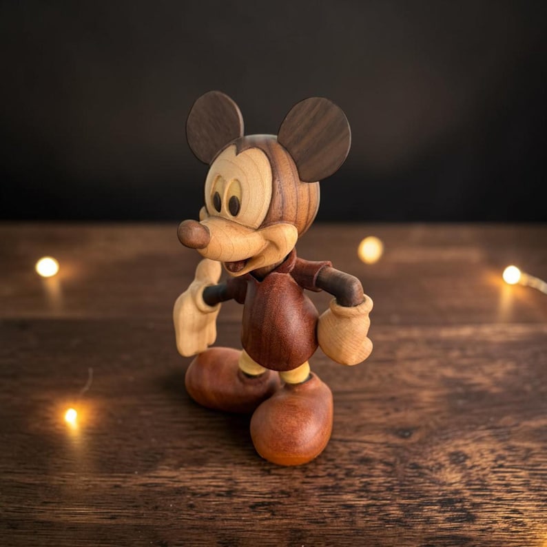 May include: A wooden Mickey Mouse figurine with a light brown face and hands, dark brown ears, and a red shirt. The figurine is standing on a wooden surface. The figure is approximately 6 inches tall.