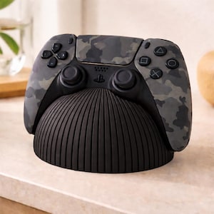 Könnte beinhalten: Ein Controller mit Tarnmuster liegt auf einer schwarzen Ladestation. Der Controller hat ein grau-, schwarz- und grünes Tarnmuster. Der Ständer hat eine abgerundete, gerippte Form. Der Controller hat schwarze Tasten und Richtungspads.