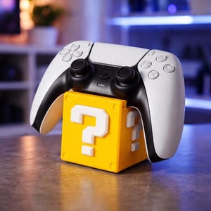Könnte beinhalten: Ein weiß-schwarzer PlayStation 5-Controller liegt auf einem gelben Block mit weißen Fragezeichen. Der Controller hat schwarze Joysticks und Tasten. Der Block ist leuchtend gelb.