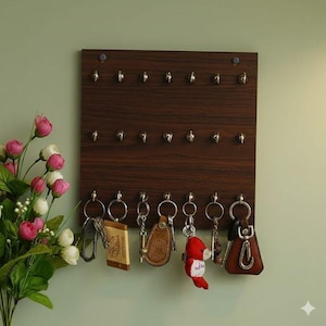 Könnte beinhalten: Ein dunkelbrauner Schlüsselhalter aus Holz mit mehreren silbernen Haken. Mehrere Schlüssel mit Schlüsselanhängern hängen an den Haken. Der Schlüsselhalter ist an einer hellgrünen Wand befestigt. Ein Strauß rosa und weißer Blumen befindet sich in der Ecke.